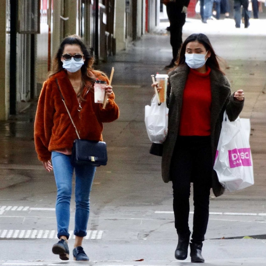 Two women walking, holding takeaway drinks and wearing face masks.