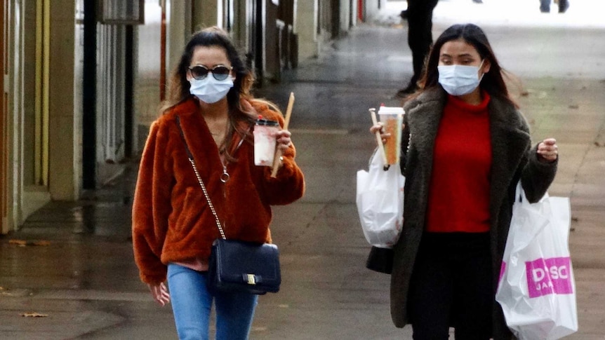 Two women walking, holding takeaway drinks and wearing face masks.