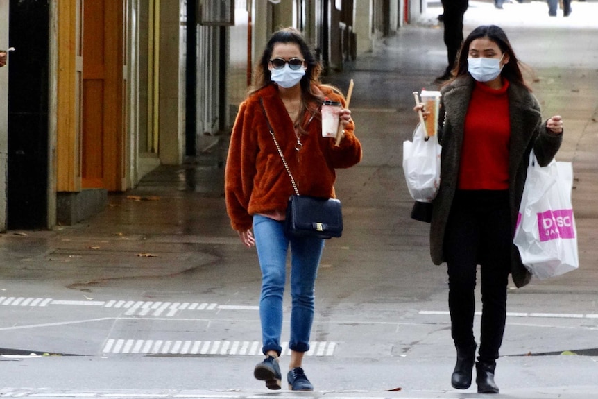 Two women walking, holding takeaway drinks and wearing face masks.