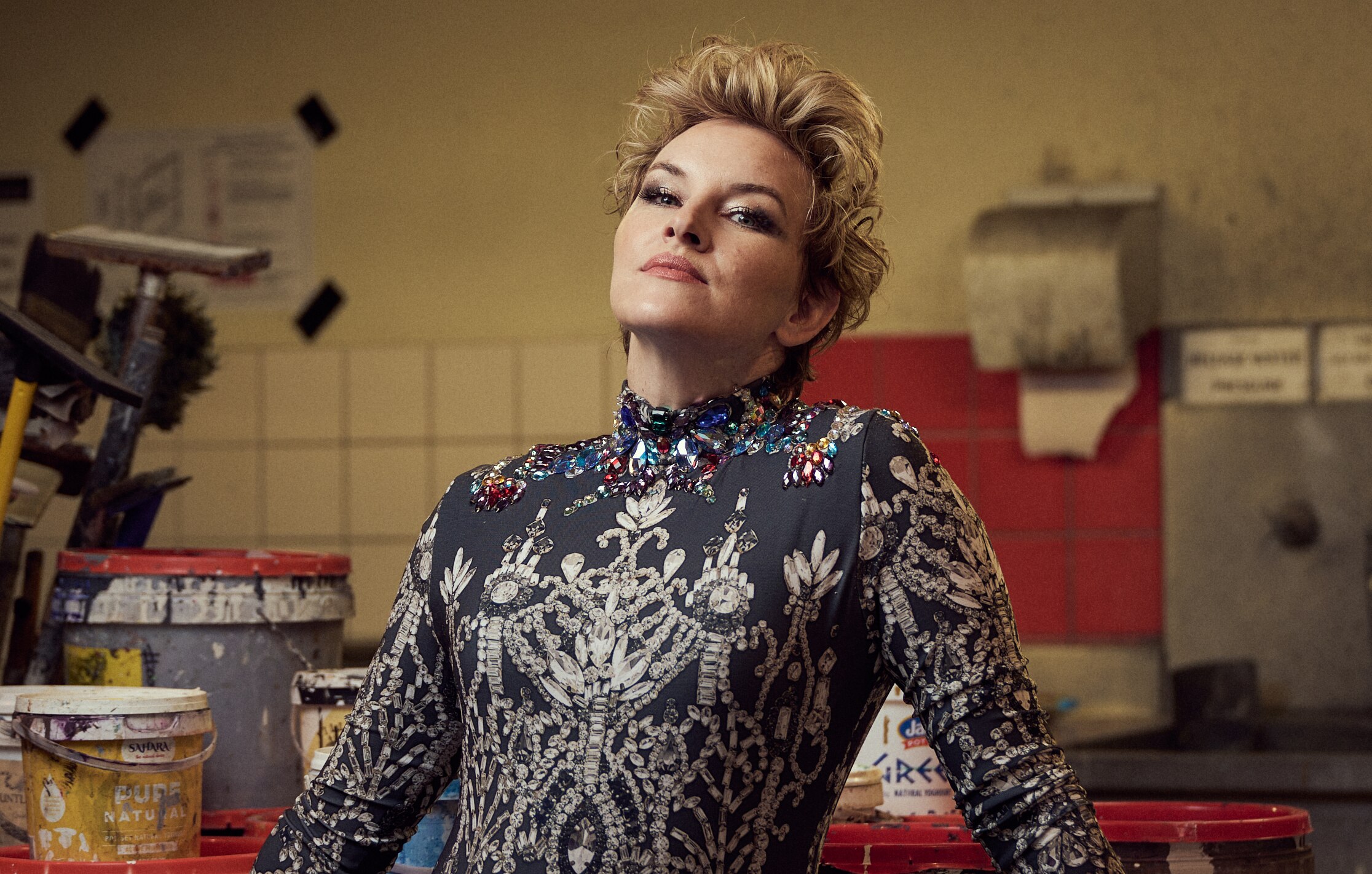 A blonde, short-haired woman in her 40s, wearing an extravagant dress, stands in a workshop, with a defiant expression