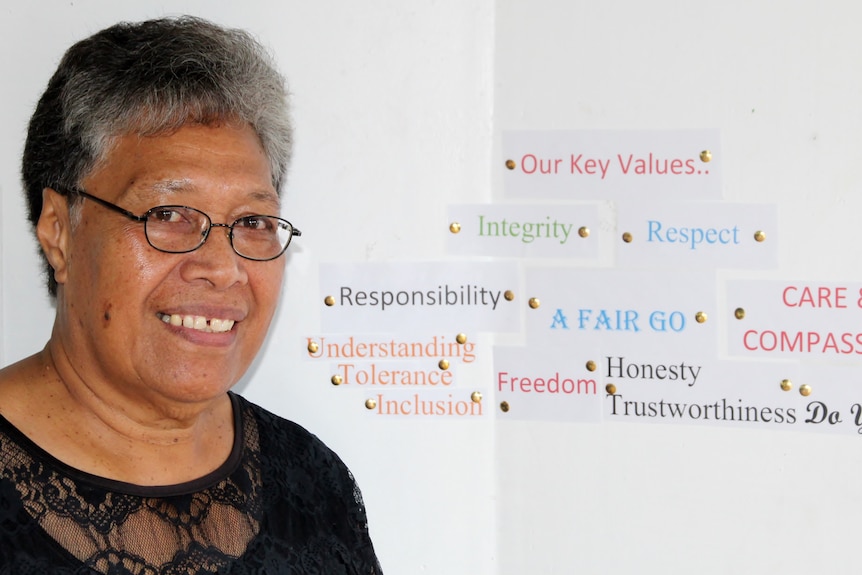 A woman with glasses stands in front of a wall with various words such as "integrity", "respect" and "a fair go".