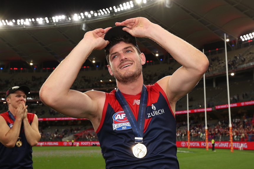 A smiling Melbourne AFL player wears a cap and has a medal around his neck after his team has won a grand final. 