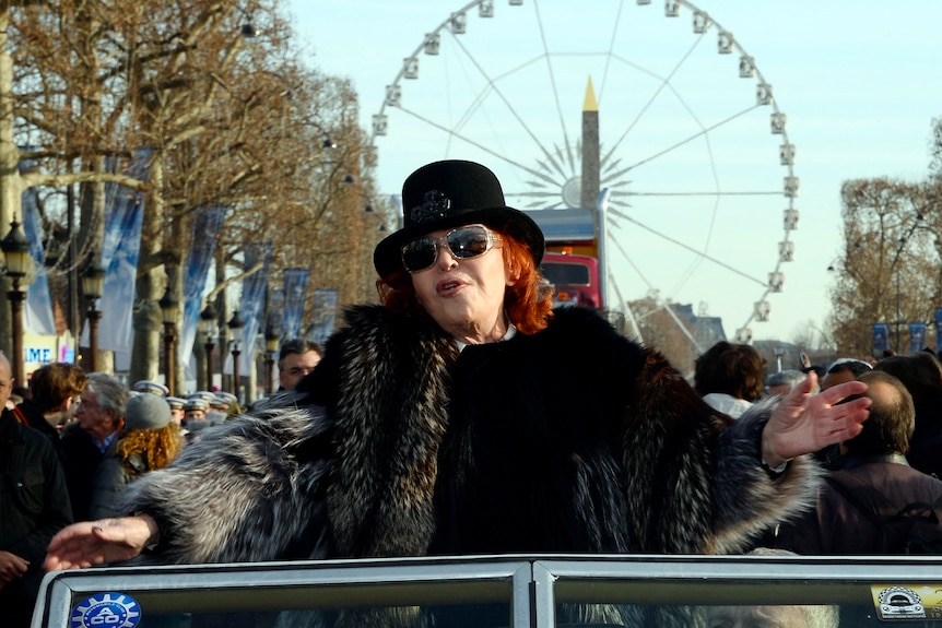 A woman dressed in a fancy hat, large sunglasses and a fur coat stands in the back seat of a convertible on a crowded street