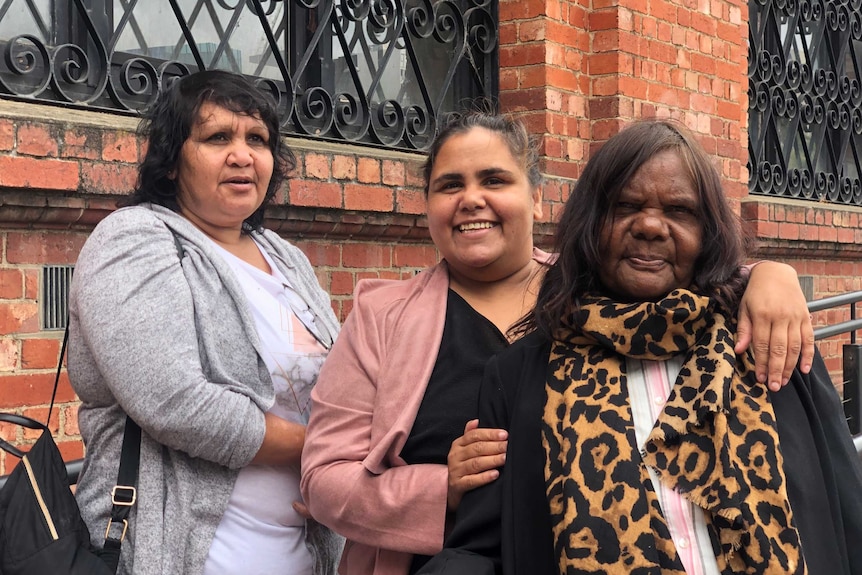 Three Aboriginal women stand in front of a red brick building