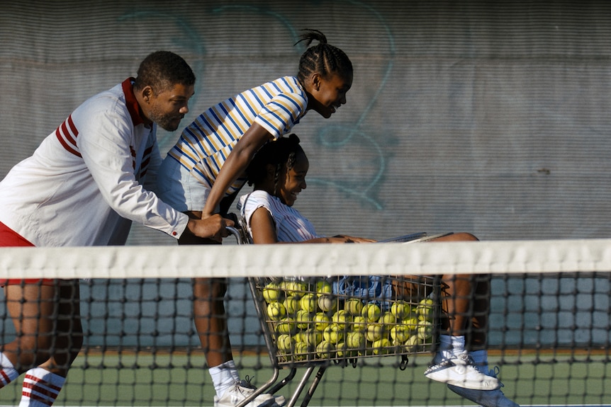 An African American man pushes two African American teenage girls along in a trolley full of a tennis balls