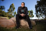 Wesley Enoch, sitting on a rock in a park on a sunny day, looks at the camera with his hands clasped.