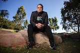 Wesley Enoch, sitting on a rock in a park on a sunny day, looks at the camera with his hands clasped.