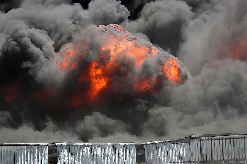 A close-up shot of thick black smoke and flames rising into the sky above a large shed roof.