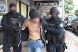 A shirtless man with a blurred face walks down a street flanked by two police officers in navy clothes and balaclavas.