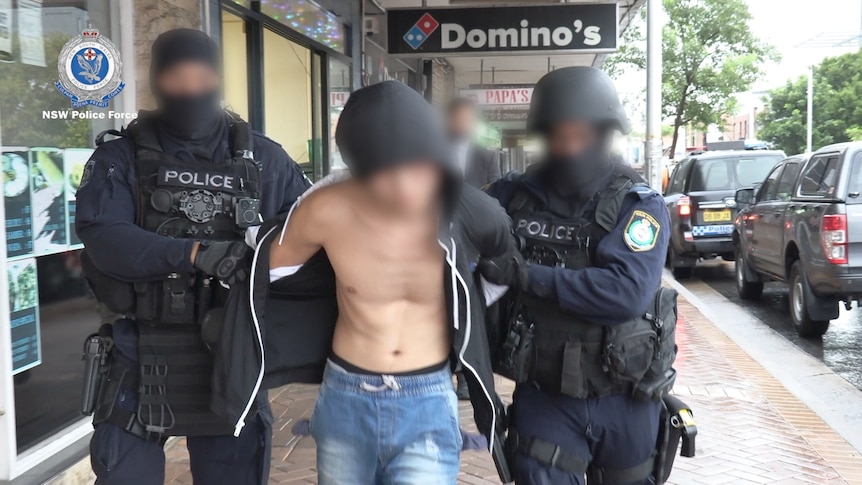 A shirtless man with a blurred face walks down a street flanked by two police officers in navy clothes and balaclavas.