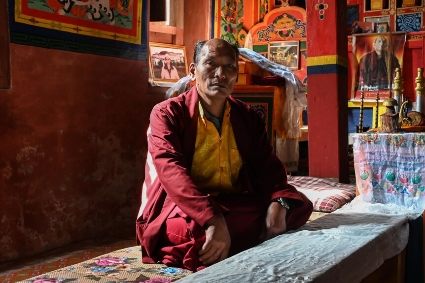 A man in maroon and yellow buddhist robes sits cross legged in a temple