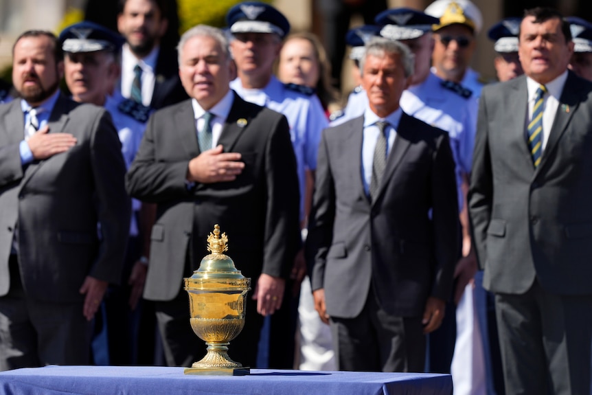 Men in suits stand in the sun with their hands on their hearts while singing and looking at a golden urn.