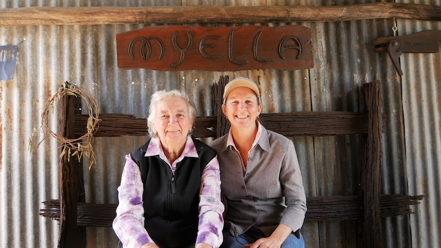 Two women, mother and adult daughter, sit in front of a corrugated iron wall. They are smiling
