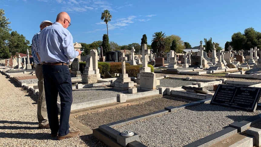 Two men stand next to each other looking down at a headstone at a cemetery