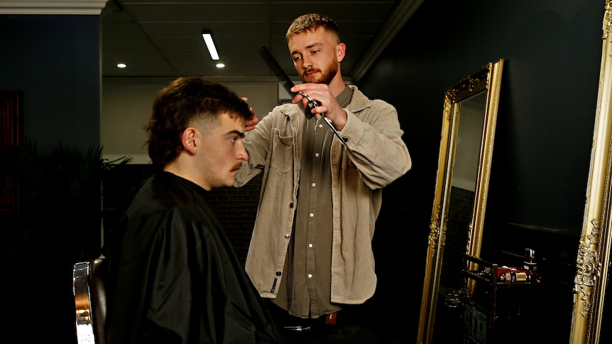 A young man holding a comb and scissors, cutting the hair of another young man seated in a chair in front of a mirror.