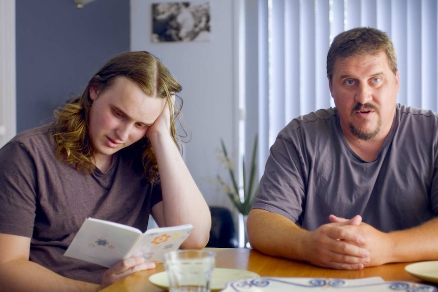 A young man with long hair reads a card at a table with his head in his hand with his father next to him