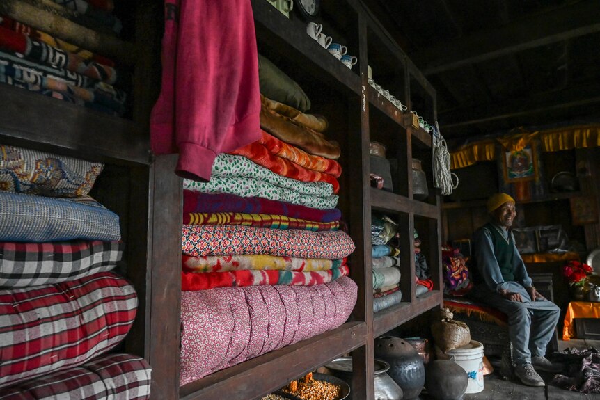 A man site on a chair in a room with colourful blankets stacked on a shelf