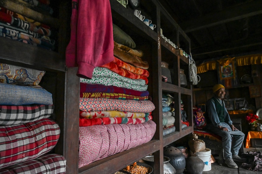 A man site on a chair in a room with colourful blankets stacked on a shelf