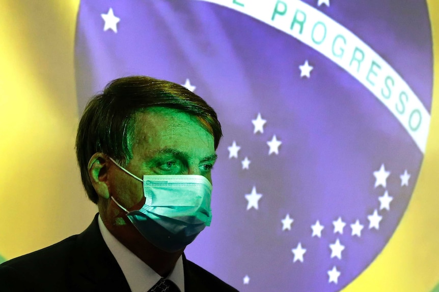 Președintele brazilian Jair Bolsonaro poartă o mască de protecție în fața steagului brazilian.