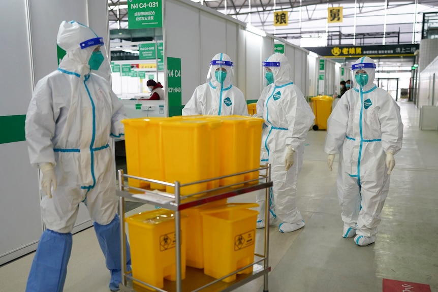 Medical workers in protective suits move a stack of yellow biohazzard containers through a large corridor.