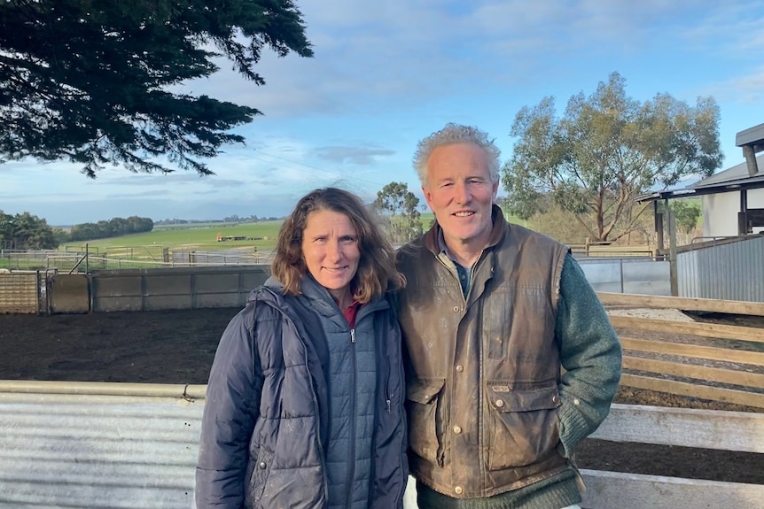 A woman and man dressed in warm clothing stand shoulder to shoulder on a farm with fenced yards behind them