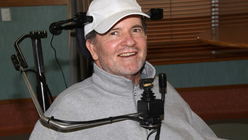 Mick McKay wearing a white cap and grey jumper sitting in a wheelchair.