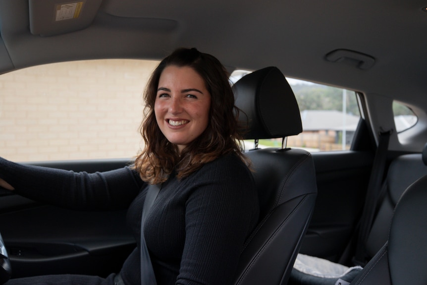 A young woman sits in the driver's seat of a car and smiles