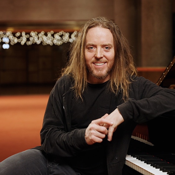 Tim Minchin sits at a grand piano. His elbow is propped on the lid and he smiles at the camera.