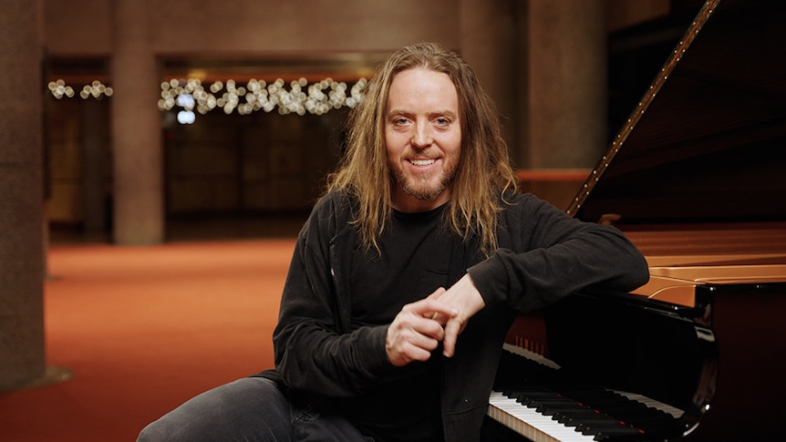 Tim Minchin sits at a grand piano. His elbow is propped on the lid and he smiles at the camera.