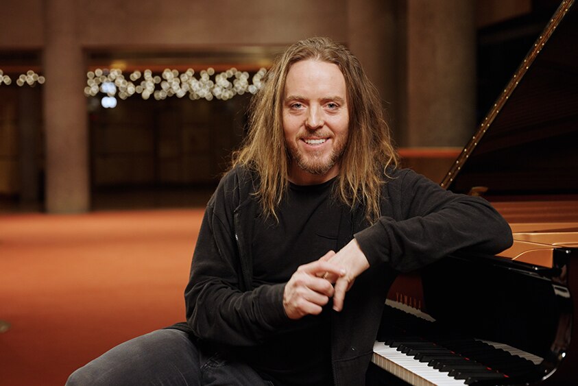 Tim Minchin sits at a grand piano. His elbow is propped on the lid and he smiles at the camera.