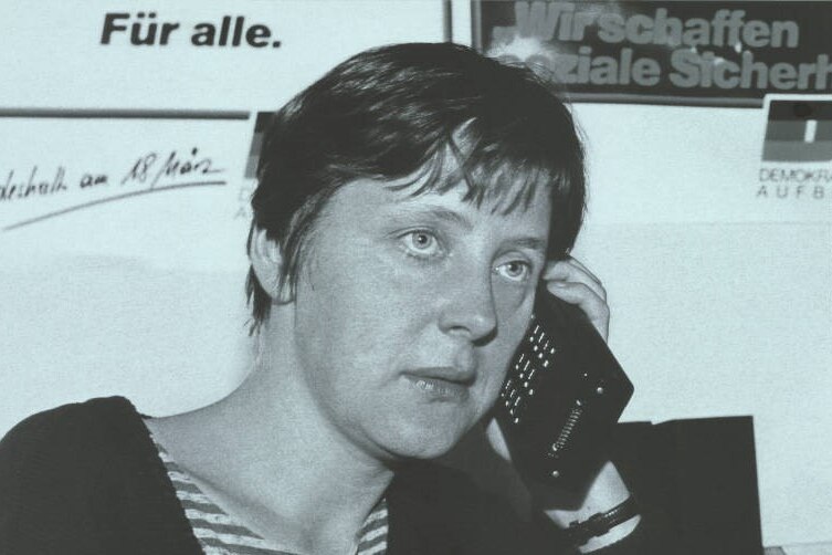 A black and white photo of a young Angela Merkel with cropped hair holding a phone to her ear