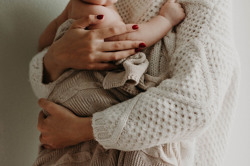 A woman with red nail polish holds a baby wrapped in a knitted blanket.