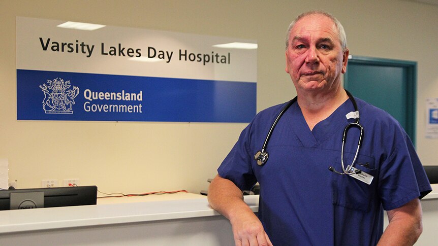 A doctor in blue scrubs stands at a hospital desk with a stethoscope around his neck