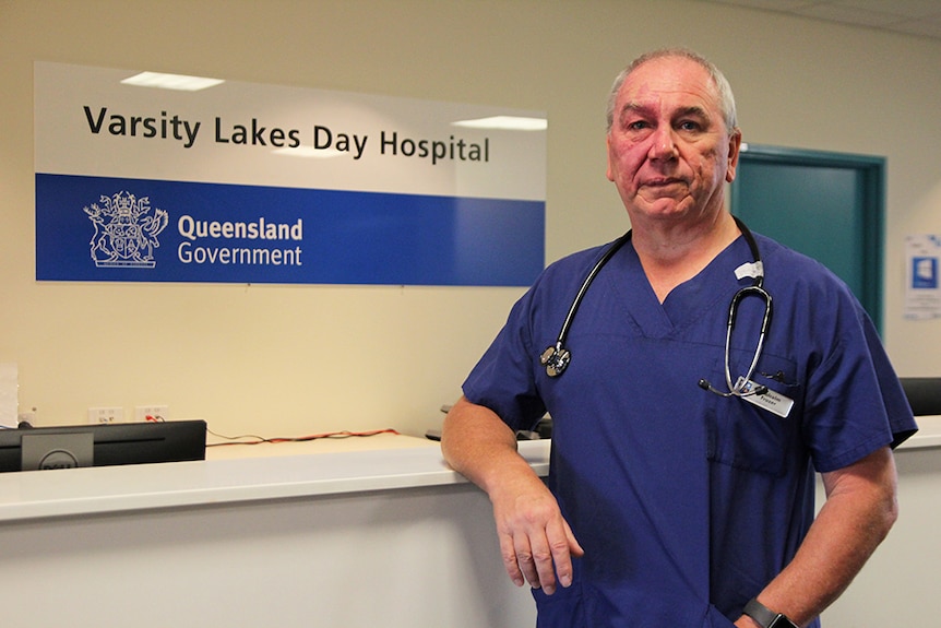 A doctor in blue scrubs stands at a hospital desk with a stethoscope around his neck