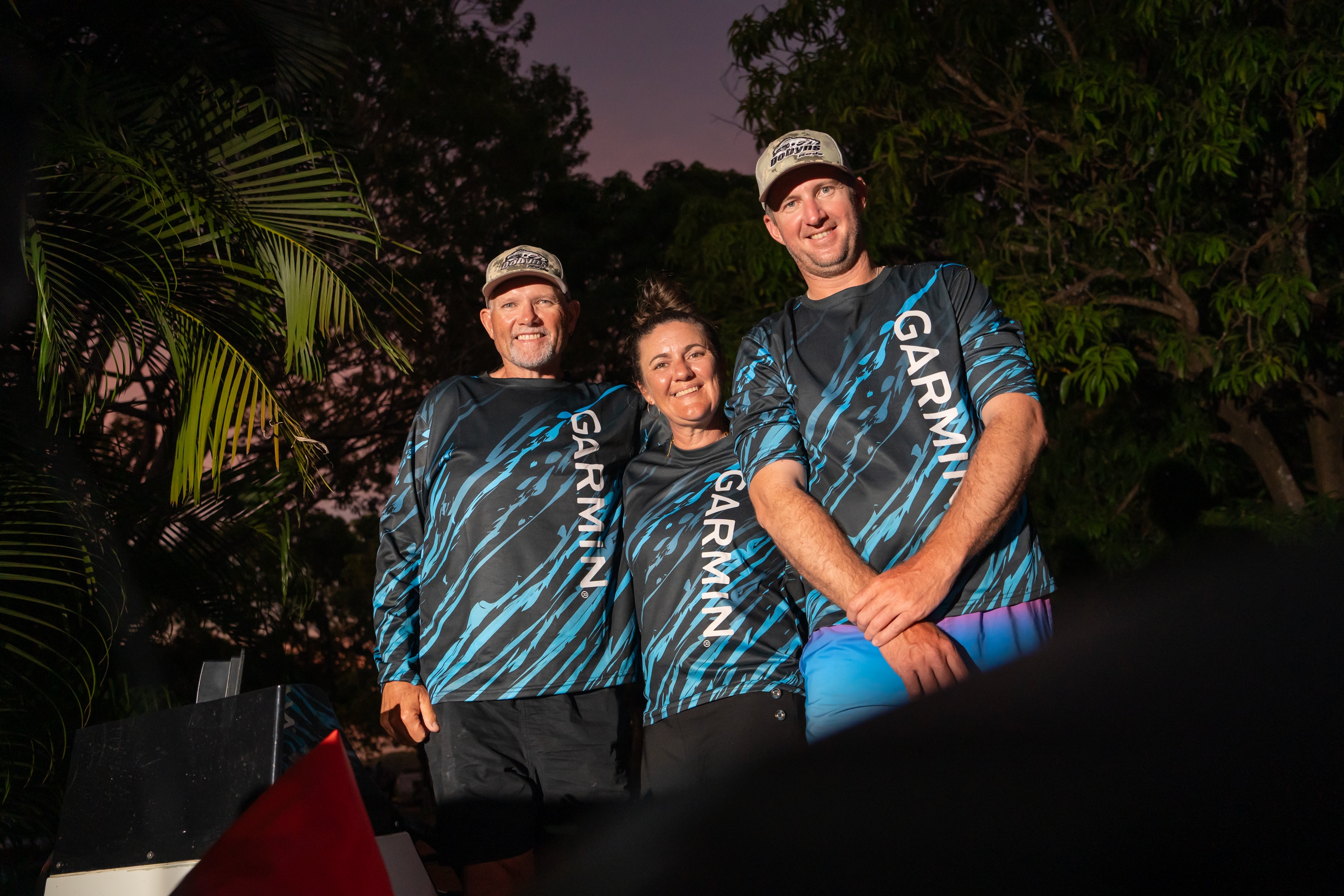 Three people (man, woman, man) standing together dark night, white light on them, all wearing blue / black longsleeve shirts