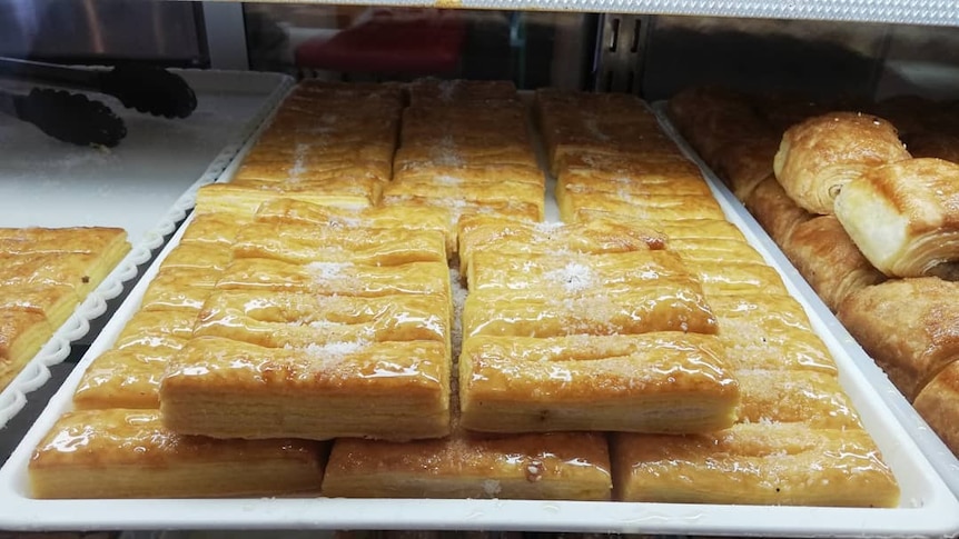 Rows of golden-glazed pastries are lined on white trays which sit under a display counter of a bakery.