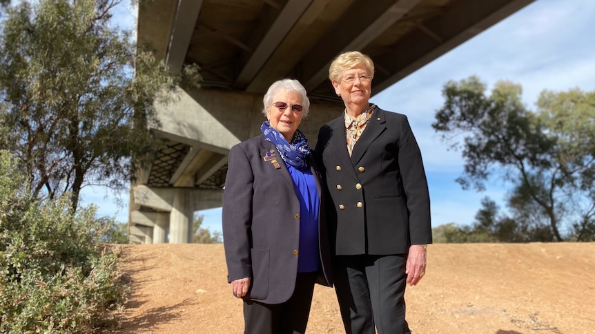 Two women stand together underneath a large bridge