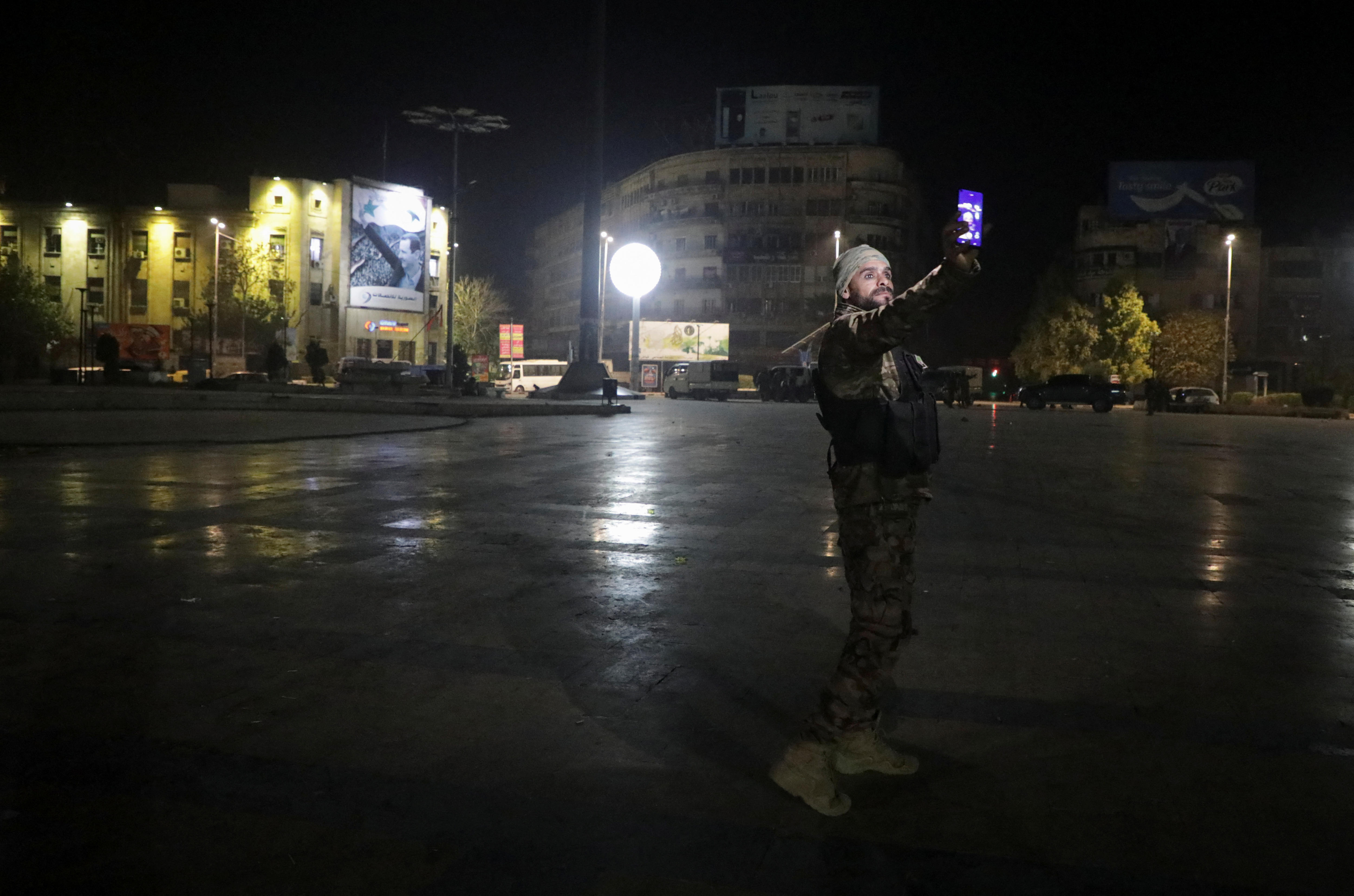 A man in camouflage combat gear takes a photo of himself with his mobile phone in a city square at night.