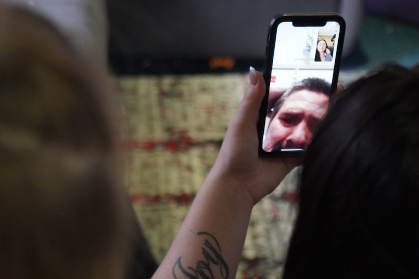A hand and tattooed arm holds up a mobile phone with a man's face showing during a video call.