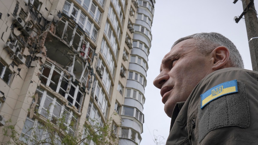 Man with Ukraine flag patch on uniform in foreground with damaged apartment building in background