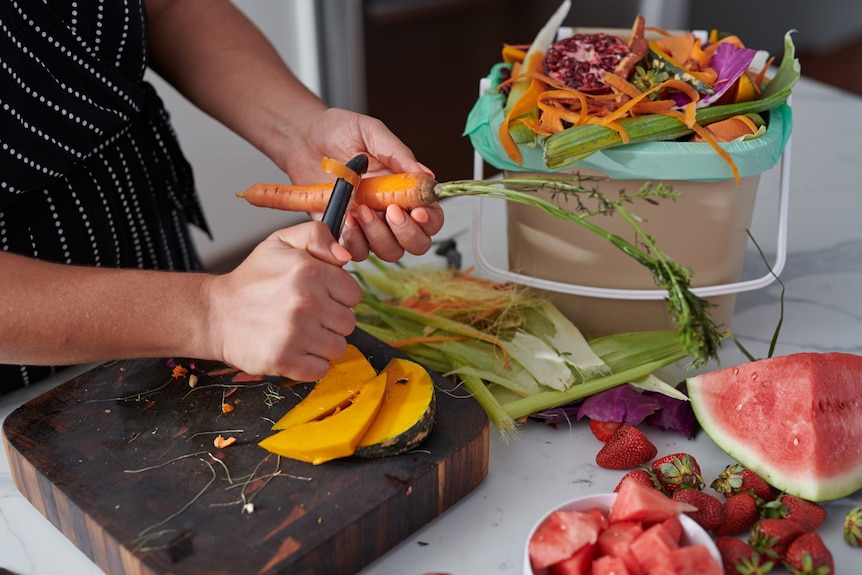 Мужчина шинкует морковь на кухонном столе перед маленькой корзиной, полной органических продуктов