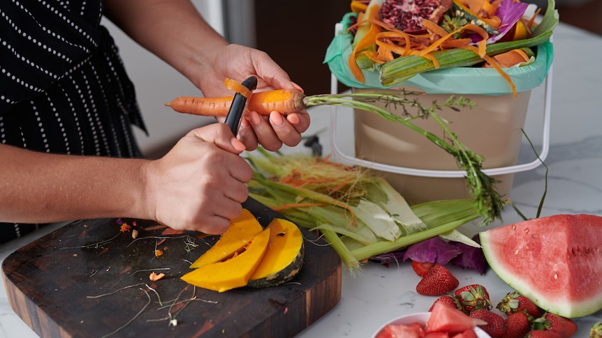 A man shreds a carrot at a kitchen bench in front of a small bin full of organic produce.
