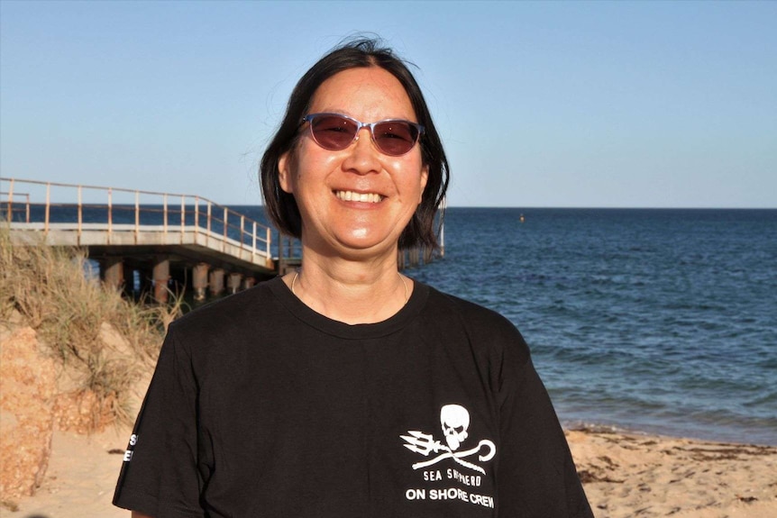 A woman in a black Sea Shepherd T-shirt and sunglasses smiles at a beach.