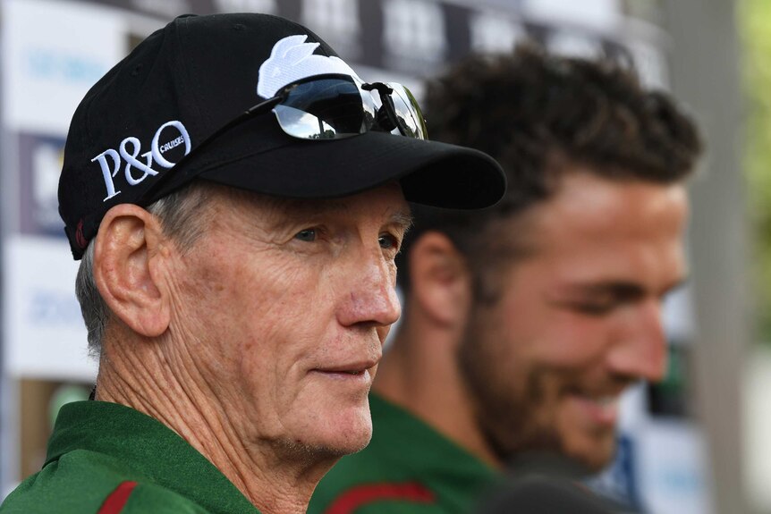Wayne Bennett, wearing a Rabbitohs cap, speaks to the media as Sam Burgess looks on in the background.