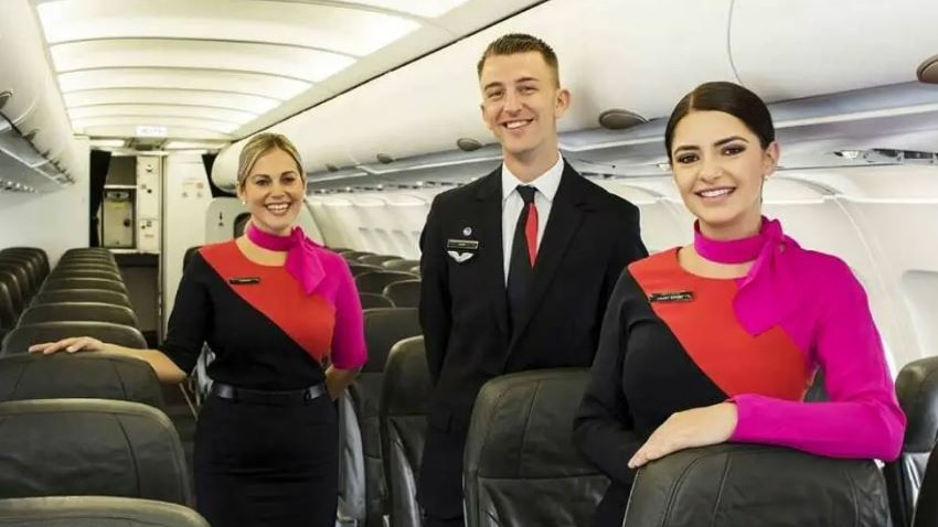 Three flight attendants — a man in a dark suit, and two women wearing red and black dresses — stand in a plane smiling.