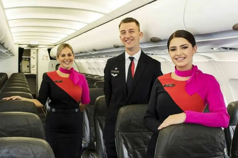 Three flight attendants — a man in a dark suit, and two women wearing red and black dresses — stand in a plane smiling.