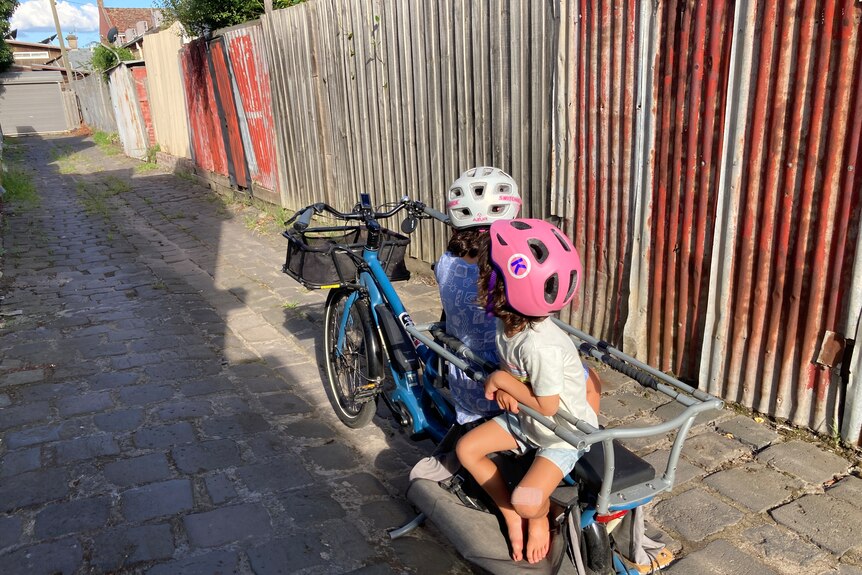 Author André Dao's electric cargo bike with two young kids sitting in a sunny driveway, an iron fence in the background.