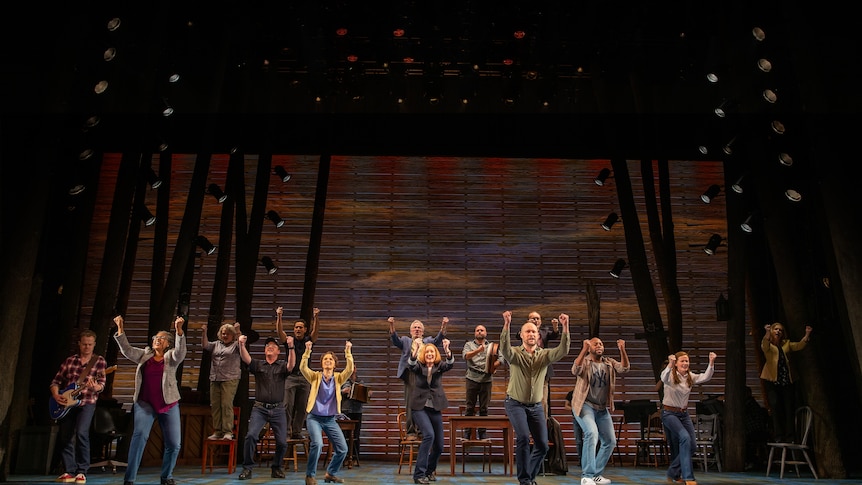 On a stage, a group of people stand with their arms up, dark tall trees in the background