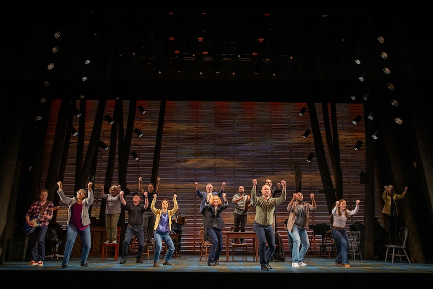 On a stage, a group of people stand with their arms up, dark tall trees in the background