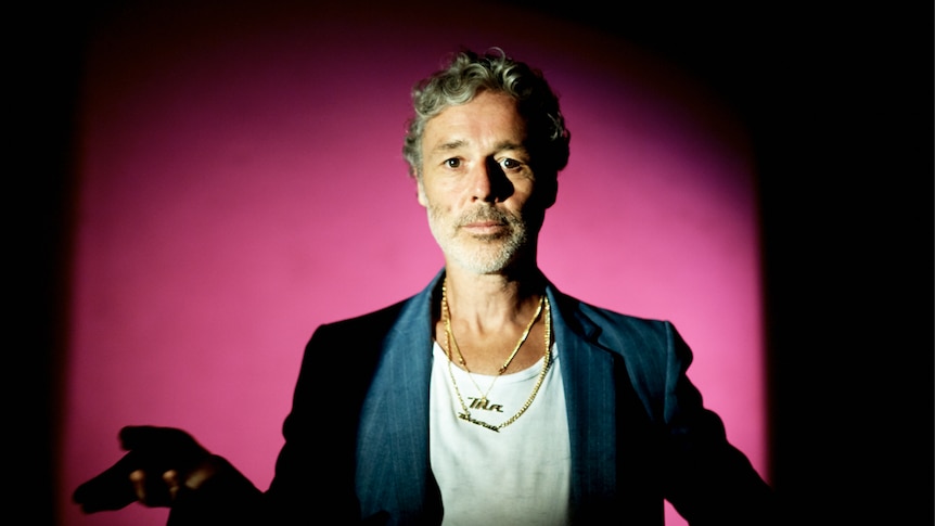 Musician Baxter Dury is photographed against a pink background. He is looking at camera wearing a blue suit jacket